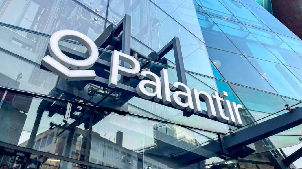 The Palantir corporate logo and name, in white 3D letters, displayed on the glass facade of a modern building.