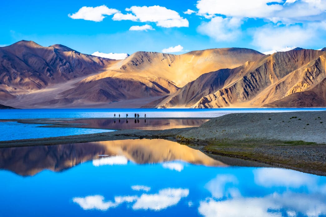 Vibrant blue Pangong Tso Lake reflecting clouds and golden mountains in Ladakh, India.