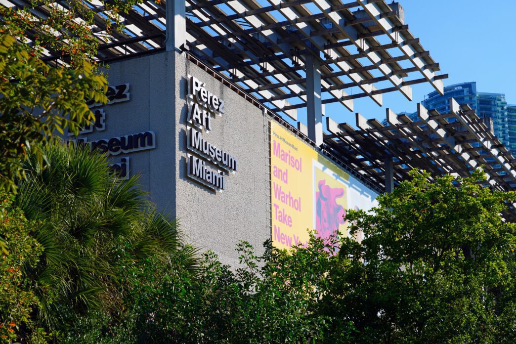 Exterior view of the Pérez Art Museum Miami (PAMM) building, showing its sign, an exhibition banner, and a wooden trellis roof structure.