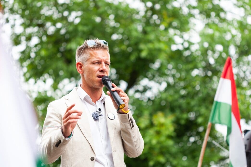 Peter Magyar, vice president of the Tisza Party, speaks into a microphone outdoors with a Hungarian flag visible