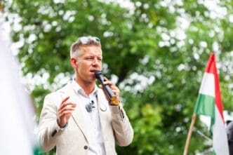 Peter Magyar, vice president of the Tisza Party, speaks into a microphone outdoors with a Hungarian flag visible