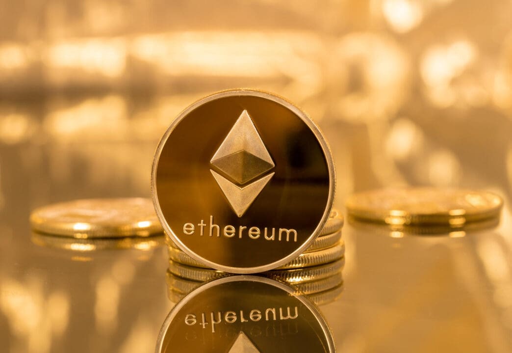 A physical Ethereum coin standing on a stack, reflected on a golden surface.