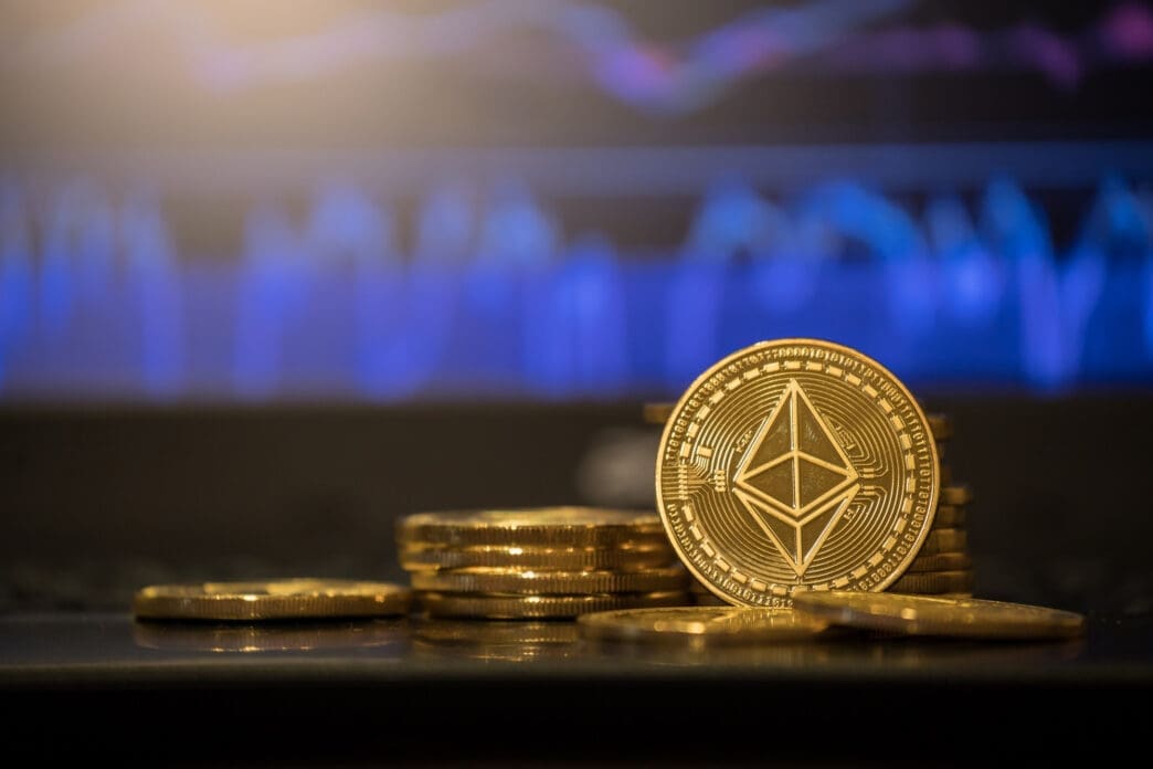 Physical gold Ethereum coins are stacked on a surface with a glowing blue financial chart blurred in the background.