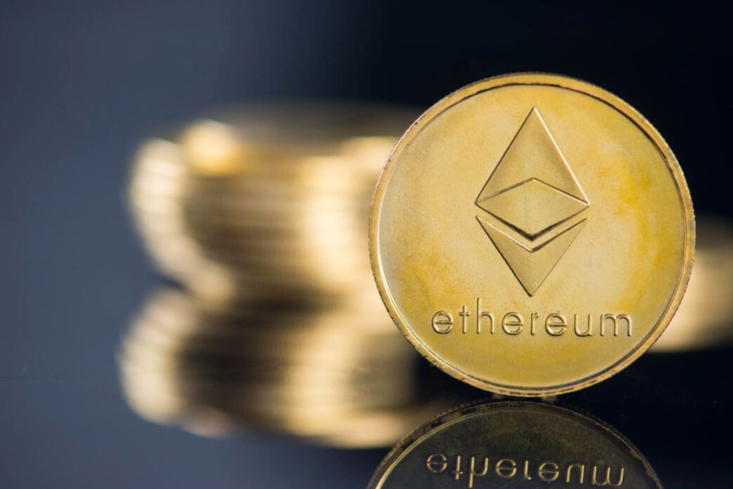 A golden Ethereum cryptocurrency coin stands upright on a reflective surface, with a stack of blurred coins behind it.
