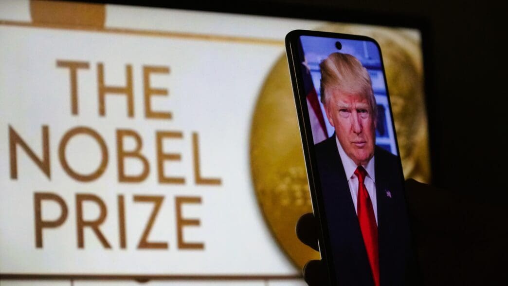 A smartphone held in a hand displaying Donald Trump's portrait with the words "The Nobel Prize" visible on a screen behind it.