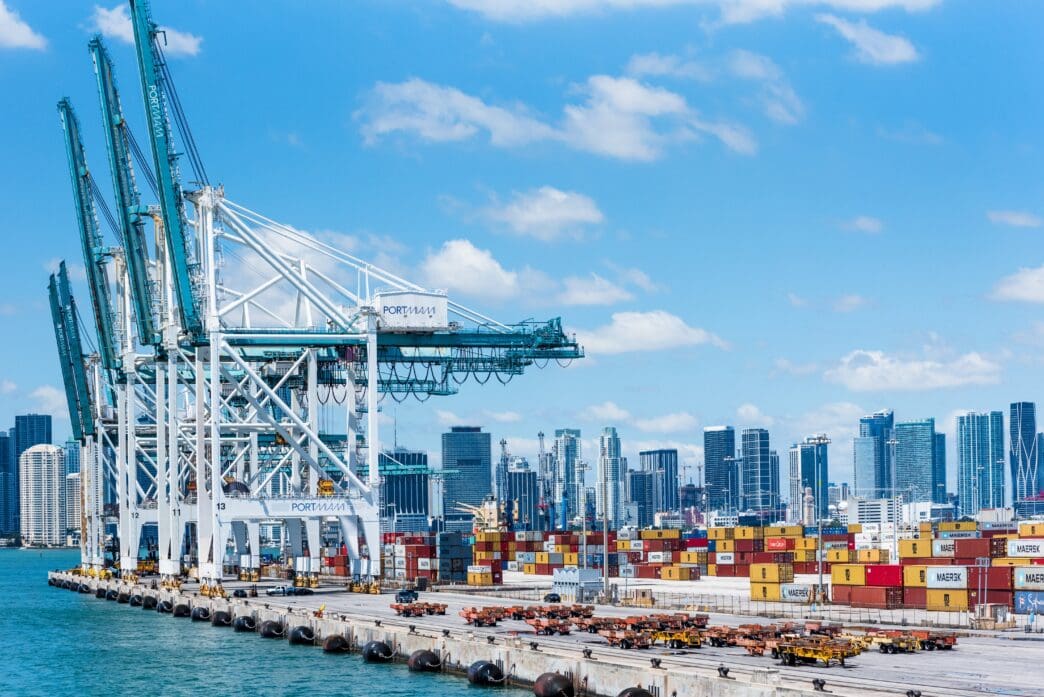Large white and teal container cranes towering over shipping containers at the Port of Miami, with the city skyline in the background.