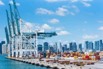 Large white and teal container cranes towering over shipping containers at the Port of Miami, with the city skyline in the background.