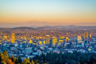 The Portland, Oregon skyline glows in golden sunset light, with the snow-capped Mount Hood visible in the distant background.