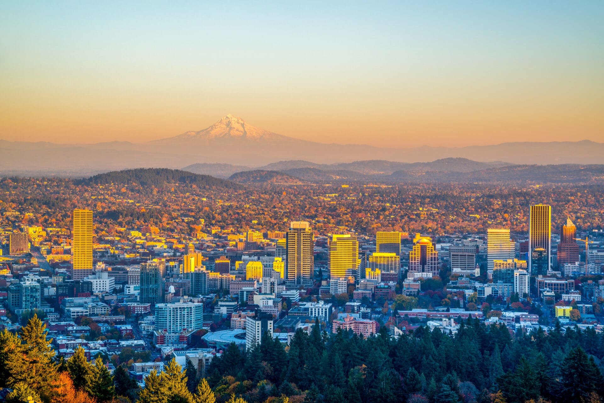 The Portland, Oregon skyline glows in golden sunset light, with the snow-capped Mount Hood visible in the distant background.