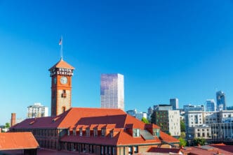The clock tower of Portland Union Station with its red roof and brick facade stands against the modern downtown skyline under a clear blue sky.