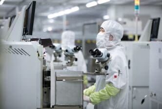 A technician in a cleanroom suit and face mask assembling electronics using a microscope at a high-tech factory