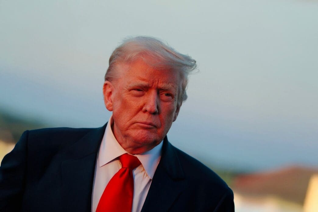 Close-up portrait of President Donald Trump in a dark suit and red tie, outdoors under a warm, reddish light.