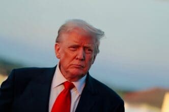 Close-up portrait of President Donald Trump in a dark suit and red tie, outdoors under a warm, reddish light.