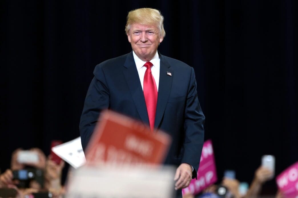 President Donald Trump smiles as he walks on stage at a rally, with supporters' signs visible in the blurred foreground.