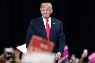President Donald Trump smiles as he walks on stage at a rally, with supporters' signs visible in the blurred foreground.