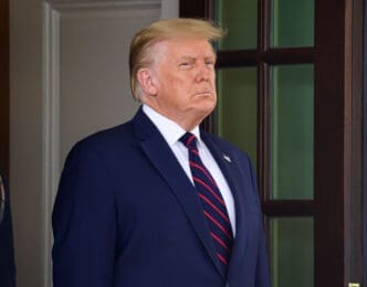 Close-up portrait of President Donald Trump in a navy suit and striped tie, standing in a doorway at the White House.