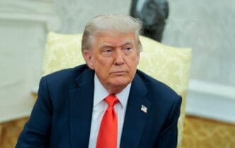 U.S. President Donald Trump, wearing a dark suit and bright red tie, sits on a gold cushioned chair looking off-camera.