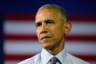 Close-up portrait of President Barack Obama with a serious, solemn expression, wearing a light blue shirt and dark patterned tie.