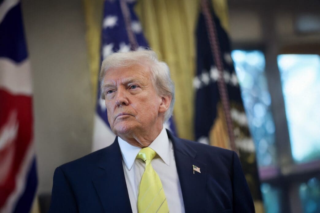 Close-up portrait of President Donald Trump in a dark suit and bright yellow tie, looking determinedly off-camera with flags in the background