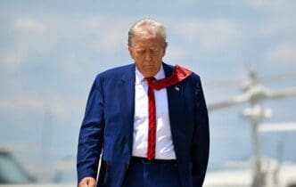 U.S. President Donald Trump, in a blue suit and red tie, walks across an airfield with a focused expression, an airplane visible behind him.