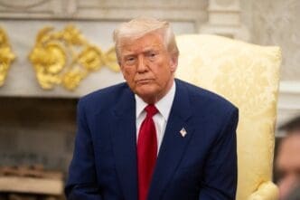 United States President Donald Trump sitting in a high-backed yellow chair, wearing a dark blue suit and bright red tie, looking directly at the camera.