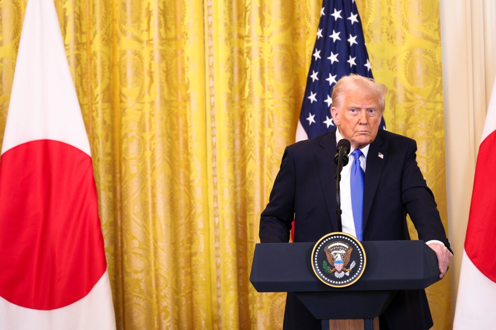 President Donald Trump stands at a podium between US and Japanese flags.