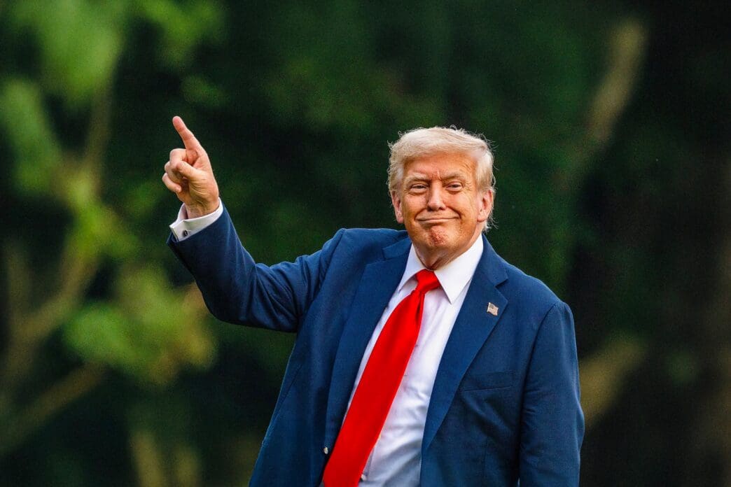 President Donald Trump points upward on the White House South Lawn, with a red tie and blue suit.