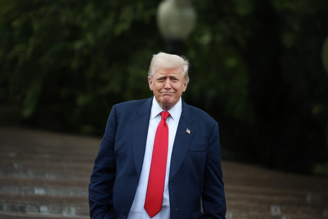 President Donald Trump, in a dark suit and red tie, walks outdoors at the White House while addressing the media.