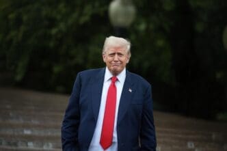 President Donald Trump, in a dark suit and red tie, walks outdoors at the White House while addressing the media.