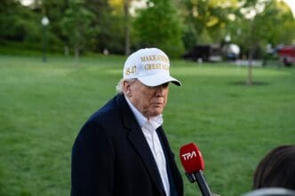 President Donald Trump, wearing a white hat with "45-47" on the side, speaks to the press on the White House lawn.