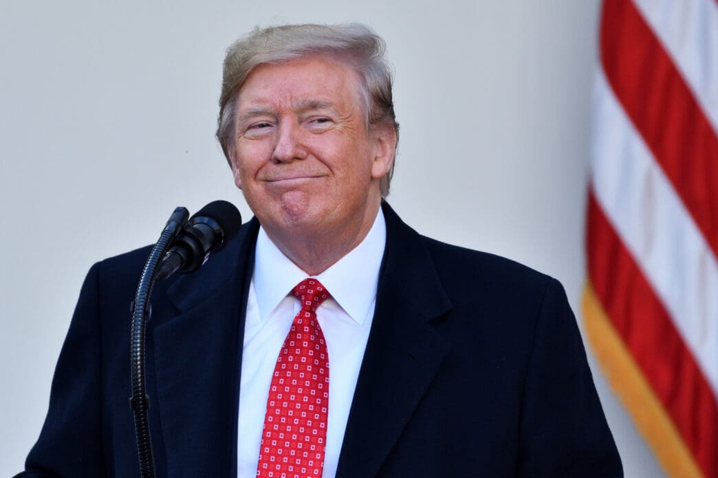 President Donald Trump smiles while speaking at a podium next to a US flag.