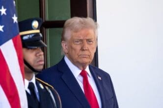 President Donald Trump, in a blue suit and red tie, standing on a bright patio next to an honor guard and the American flag