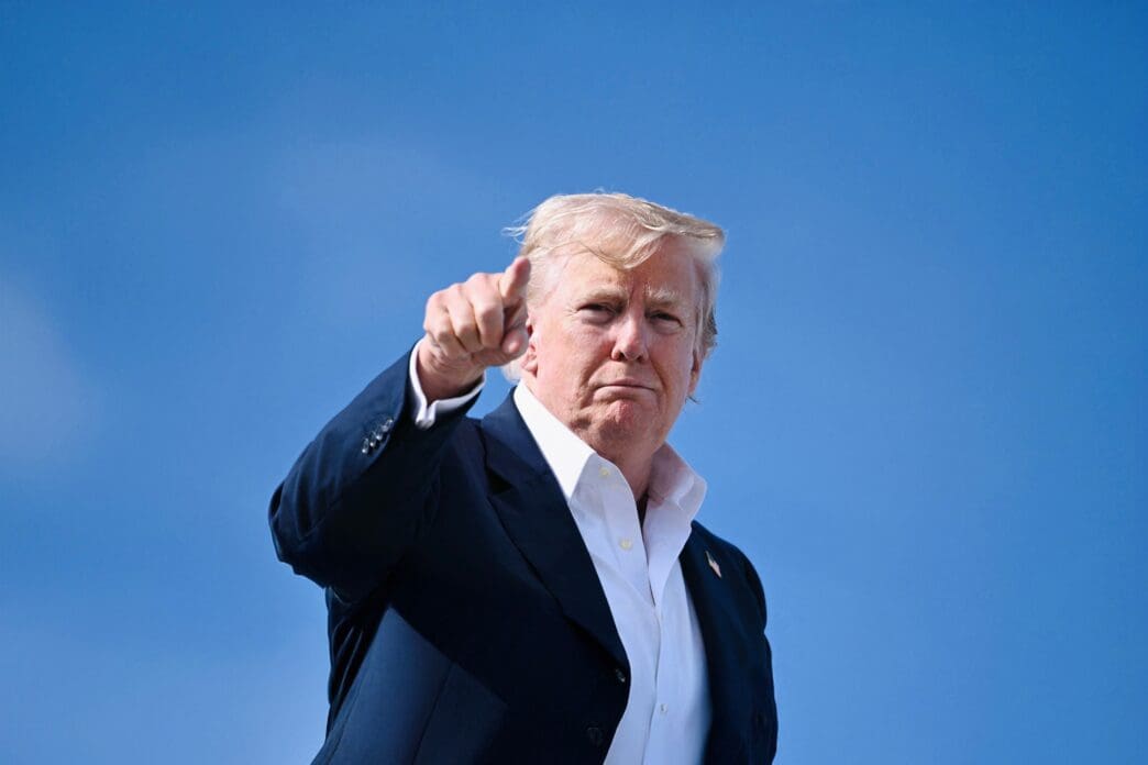 Donald Trump stands against a bright blue sky, pointing directly at the camera
