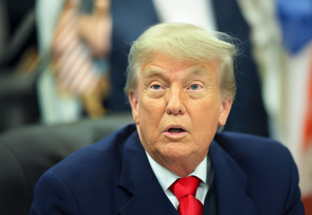 Close-up portrait of President Donald Trump in a blue suit and red tie, speaking or listening intently during a meeting.