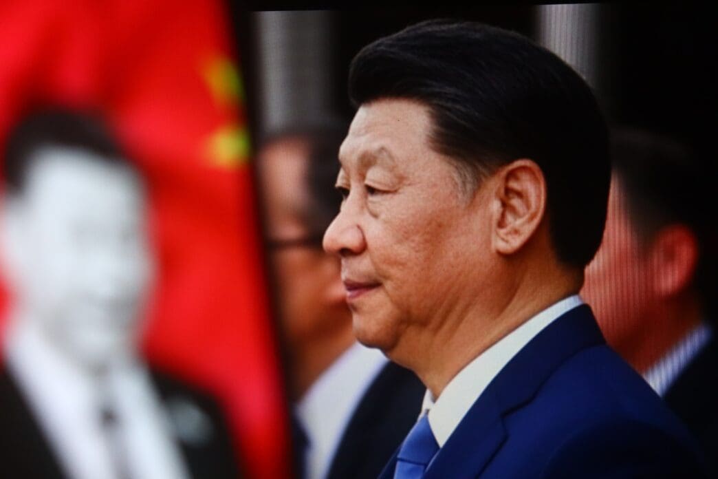 Profile close-up of President Xi Jinping in a dark suit against a blurry background with a red flag.