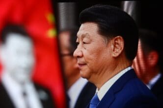 Profile close-up of President Xi Jinping in a dark suit against a blurry background with a red flag.