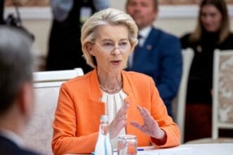 European Commission President Ursula von der Leyen speaks while wearing an orange jacket at a formal conference table.