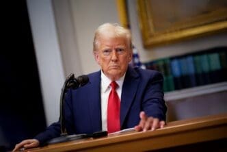 President Donald Trump, in a blue suit and red tie, seated at a table with his hand extended while addressing a microphone