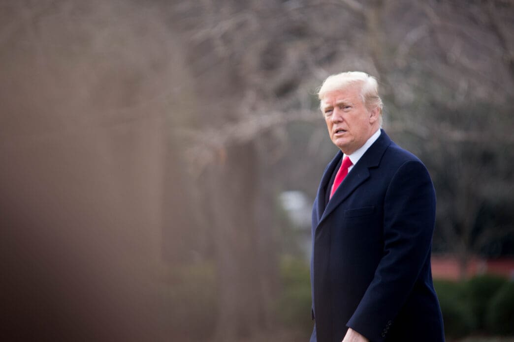 President Donald Trump in a dark overcoat and red tie walking outdoors on the White House grounds with a focused expression.