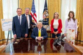 President Donald Trump seated at the Resolute Desk in the Oval Office, posing with three men and two women.