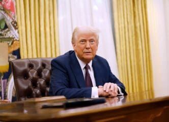 President Donald Trump seated at the Resolute Desk in the Oval Office, wearing a blue suit, with his hands clasped on the desk.