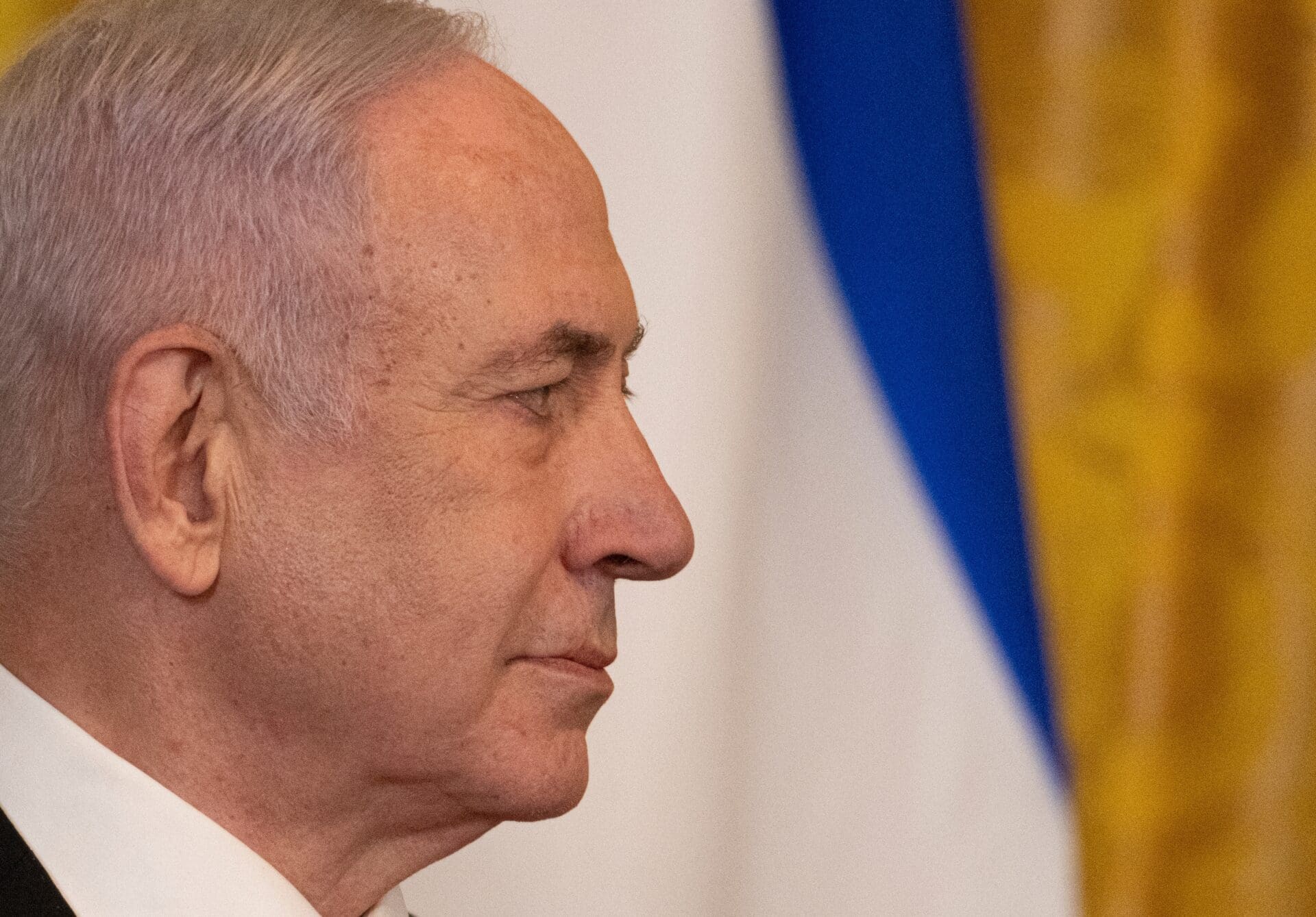 Close-up profile of Israeli Prime Minister Benjamin Netanyahu's face, with a blurred Israeli flag in the background.