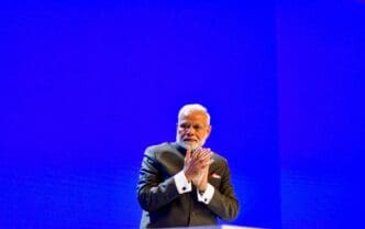 India's Prime Minister Narendra Modi stands on a stage, with hands clasped, in front of a brilliant blue screen.