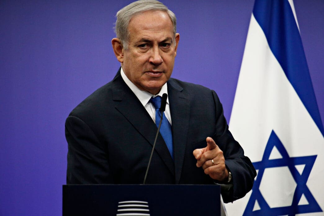 Israeli Prime Minister Benjamin Netanyahu in a dark suit points a finger while speaking at a podium with the flag of Israel to his right.