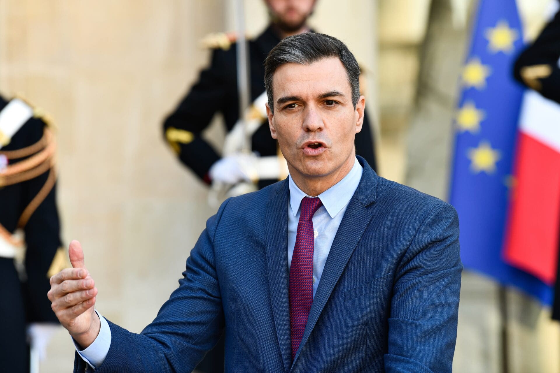 Spanish Prime Minister Pedro Sánchez gestures while speaking outdoors at the Élysée Palace, with military guards in the blurred background.