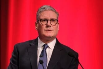 Prime Minister Sir Keir Starmer, wearing a dark suit and glasses, speaks at a podium in front of a vibrant red background.
