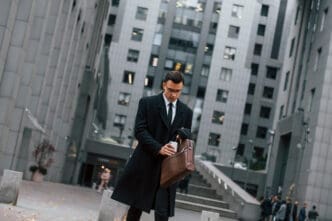 A man in a black overcoat and glasses walks in a city plaza, adjusting a brown briefcase while holding a cup, against a tall gray office building.