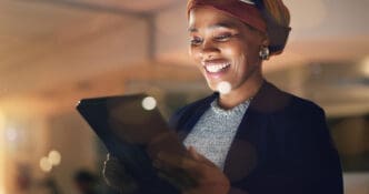 Smiling woman wearing a head wrap and a blazer holds a tablet, illuminated by its screen in a warm, low-light environment.