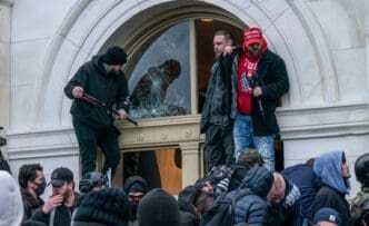 Three protesters stand at a shattered window opening of the US Capitol building entrance, with one holding a weapon-like object and another wearing a red cap.