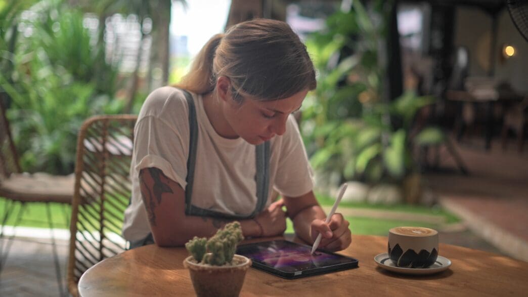 Woman with a ponytail and arm tattoos works on a digital tablet with a stylus, seated at a wooden table with a latte and a cactus.